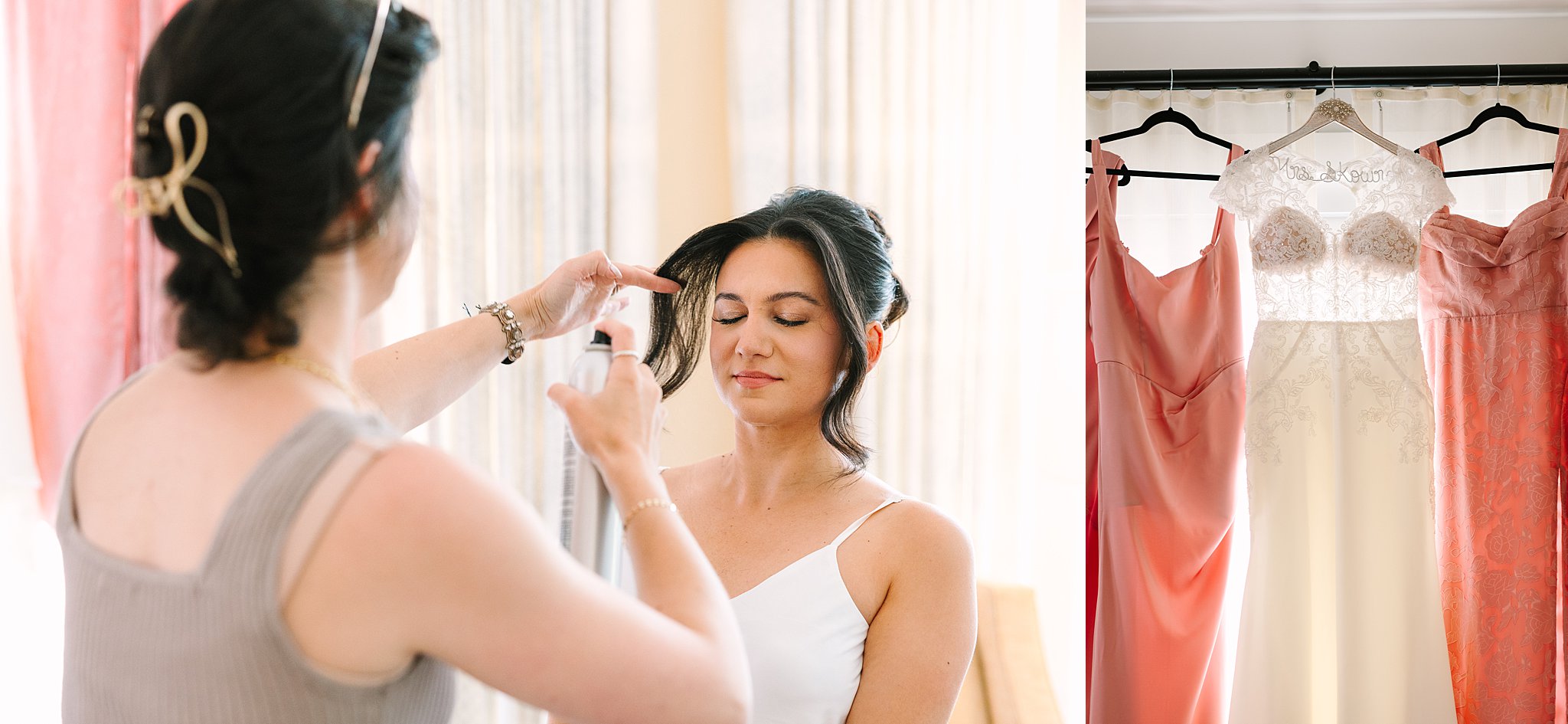 A woman with closed eyes has her hair styled with spray by another woman. Next to them, white and pink dresses hang on display, suggesting a wedding or special occasion.