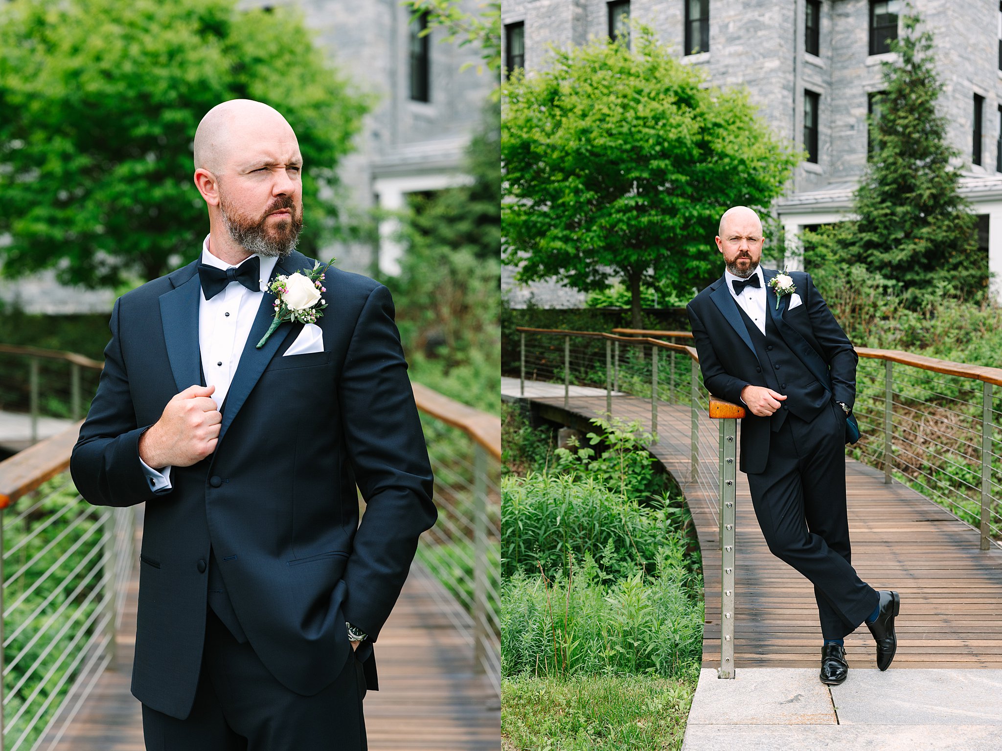A bald man with a beard in a black tuxedo and bow tie poses confidently on a wooden bridge with greenery and a modern building in the background.
