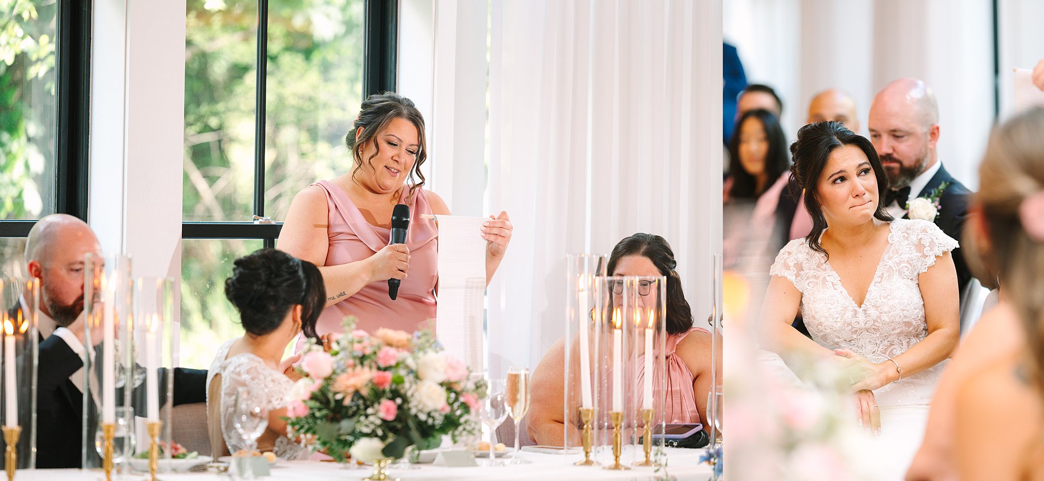 A woman in a pink dress gives a speech with a microphone at a wedding reception. The bride and other guests listen attentively at a table decorated with flowers and candles.