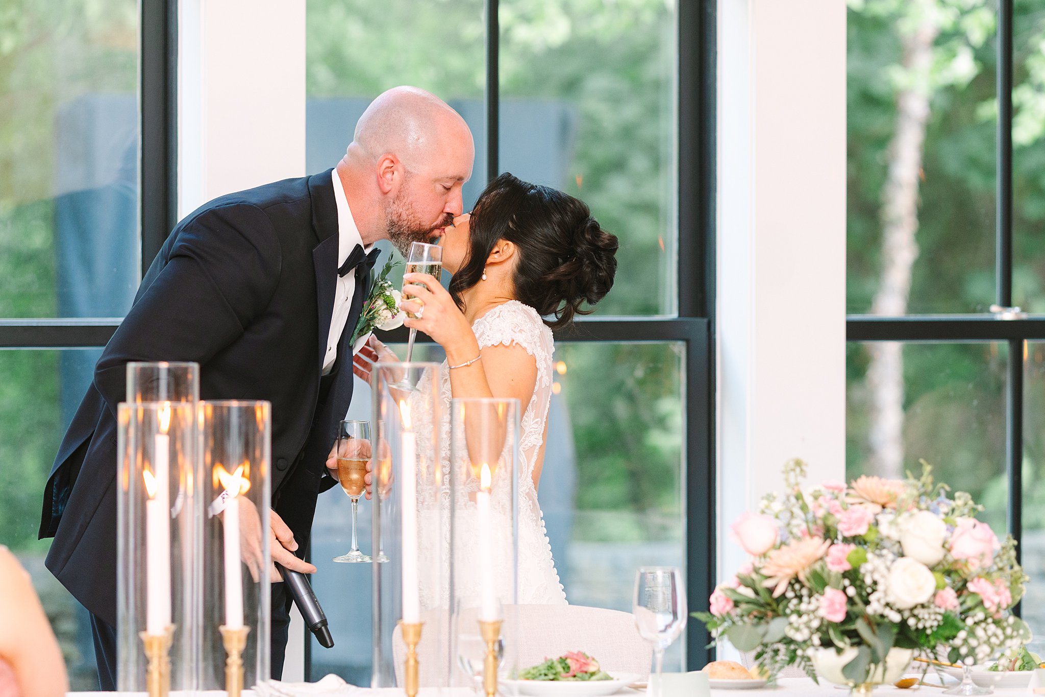 A bride and groom share a kiss during their wedding reception, holding glasses of wine. The bride wears a white dress, the groom a black tuxedo. Candles and a floral arrangement decorate the table in front of them.