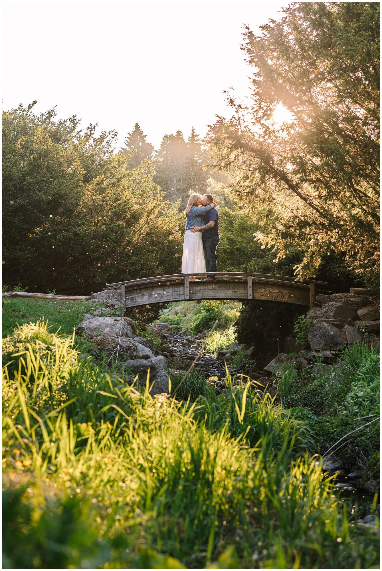 A couple embraces and kisses on a small wooden bridge surrounded by lush greenery and trees, with warm sunlight filtering through the branches in the background.