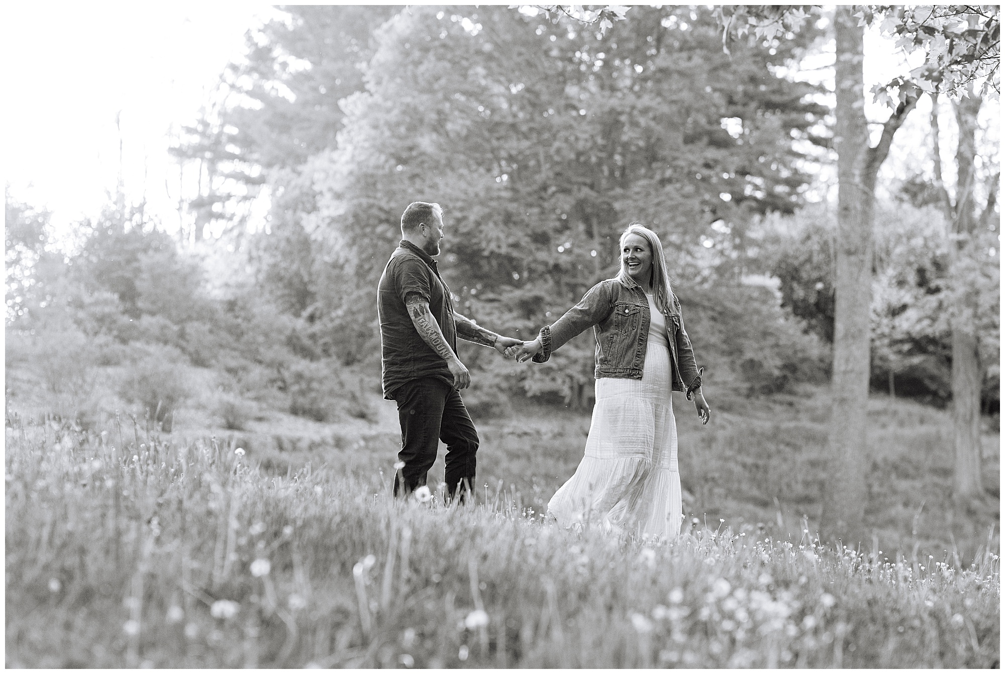 A couple holds hands and smiles at each other while walking through a grassy, flower-filled field with trees in the background. This black and white shot captures the charm of Arnold Arboretum engagement photos.
