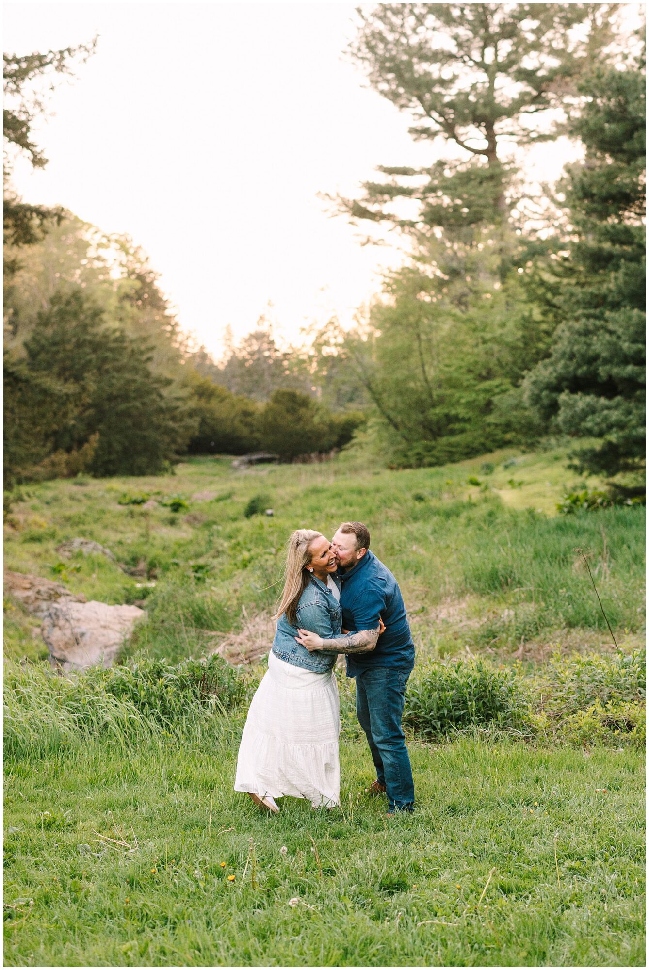 A man embraces and kisses a woman on the cheek in a grassy, green meadow with trees in the background at sunset. The woman smiles and wears a white skirt and denim jacket; the man wears jeans and a blue shirt.