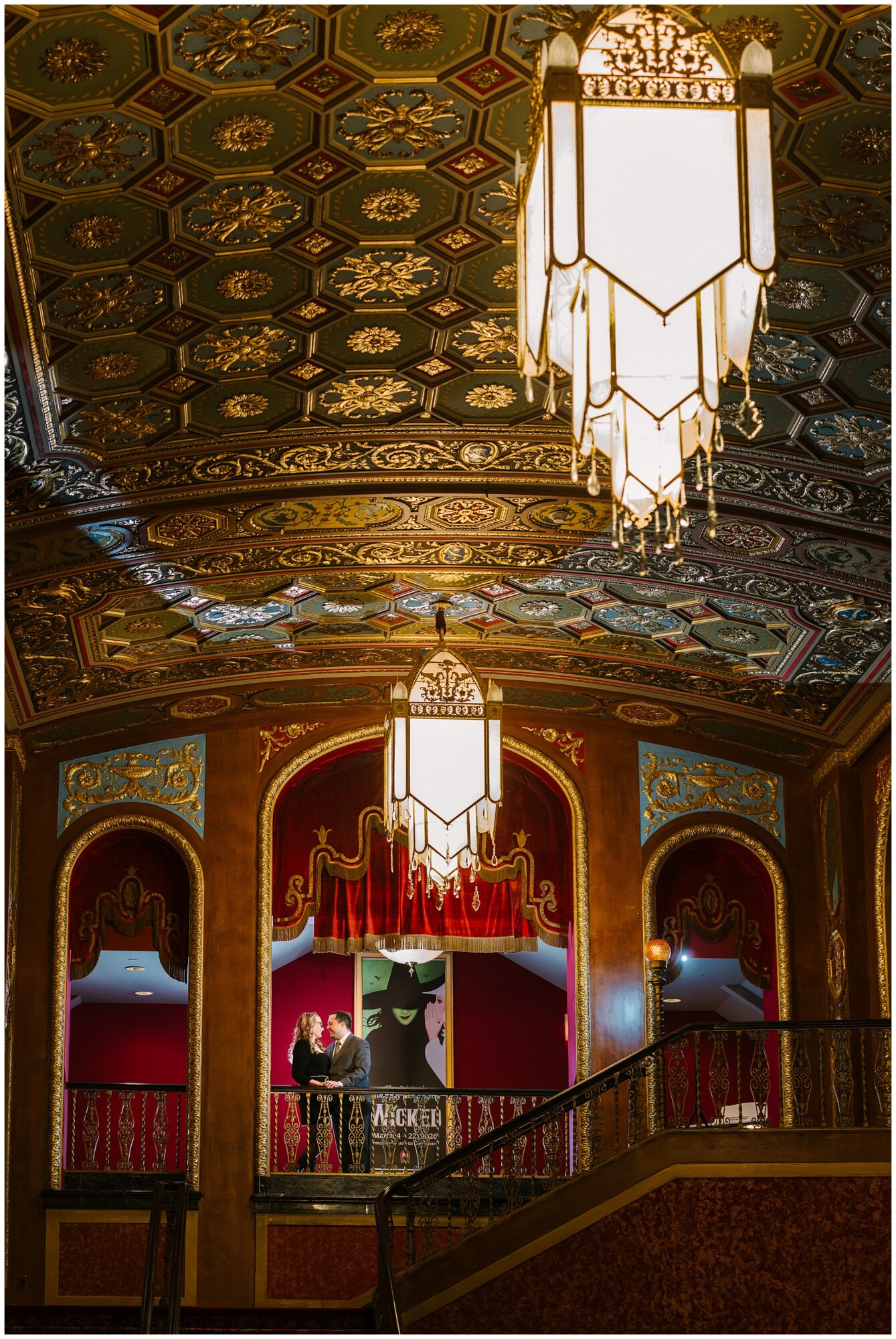 A couple stands on a grand staircase beneath ornate chandeliers and a richly decorated ceiling in an opulent theater lobby with red and gold accents.