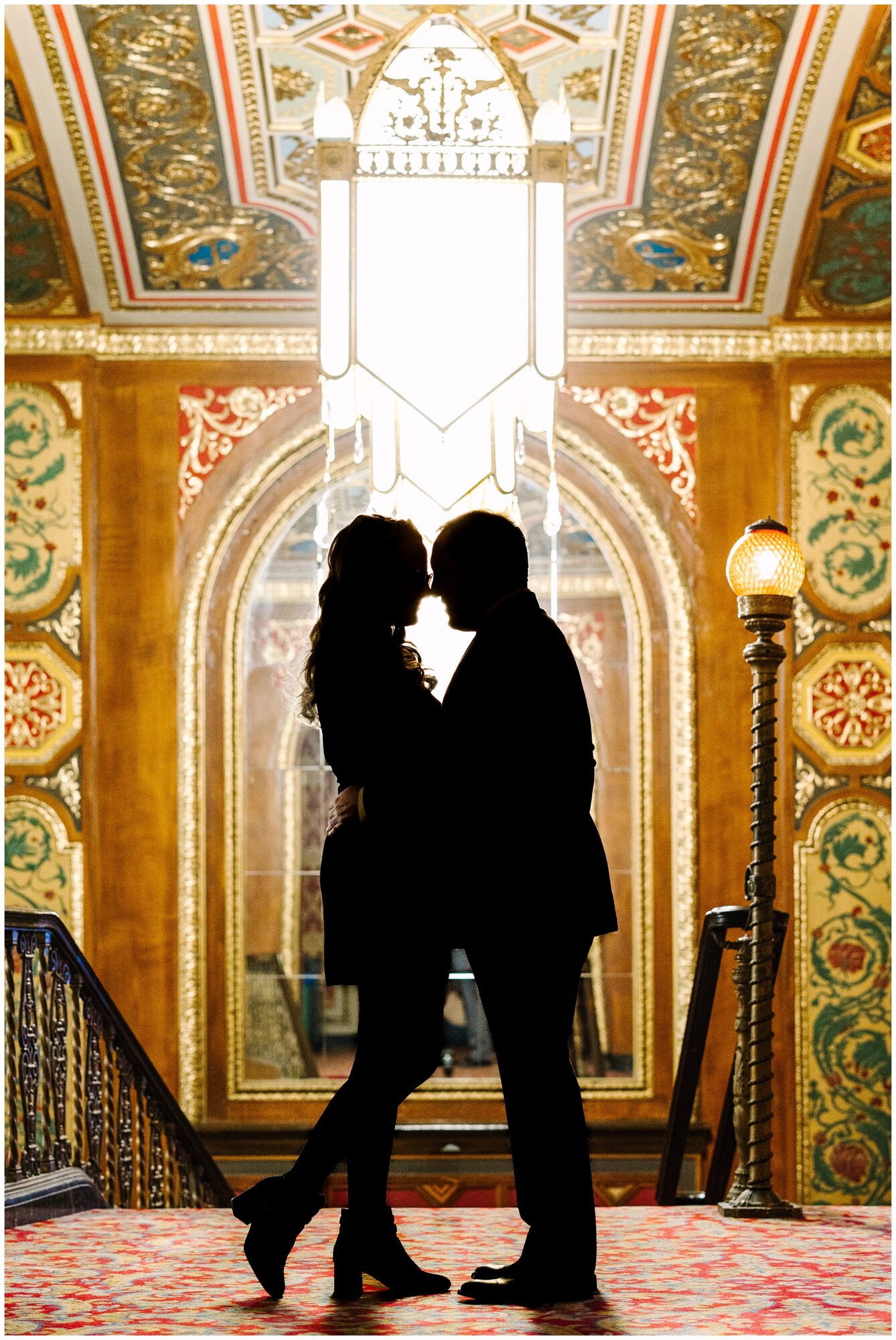 A silhouetted couple stands close together, almost kissing, in front of an ornate, golden arched window with intricate patterns and a grand chandelier above them in a lavish, historic interior.