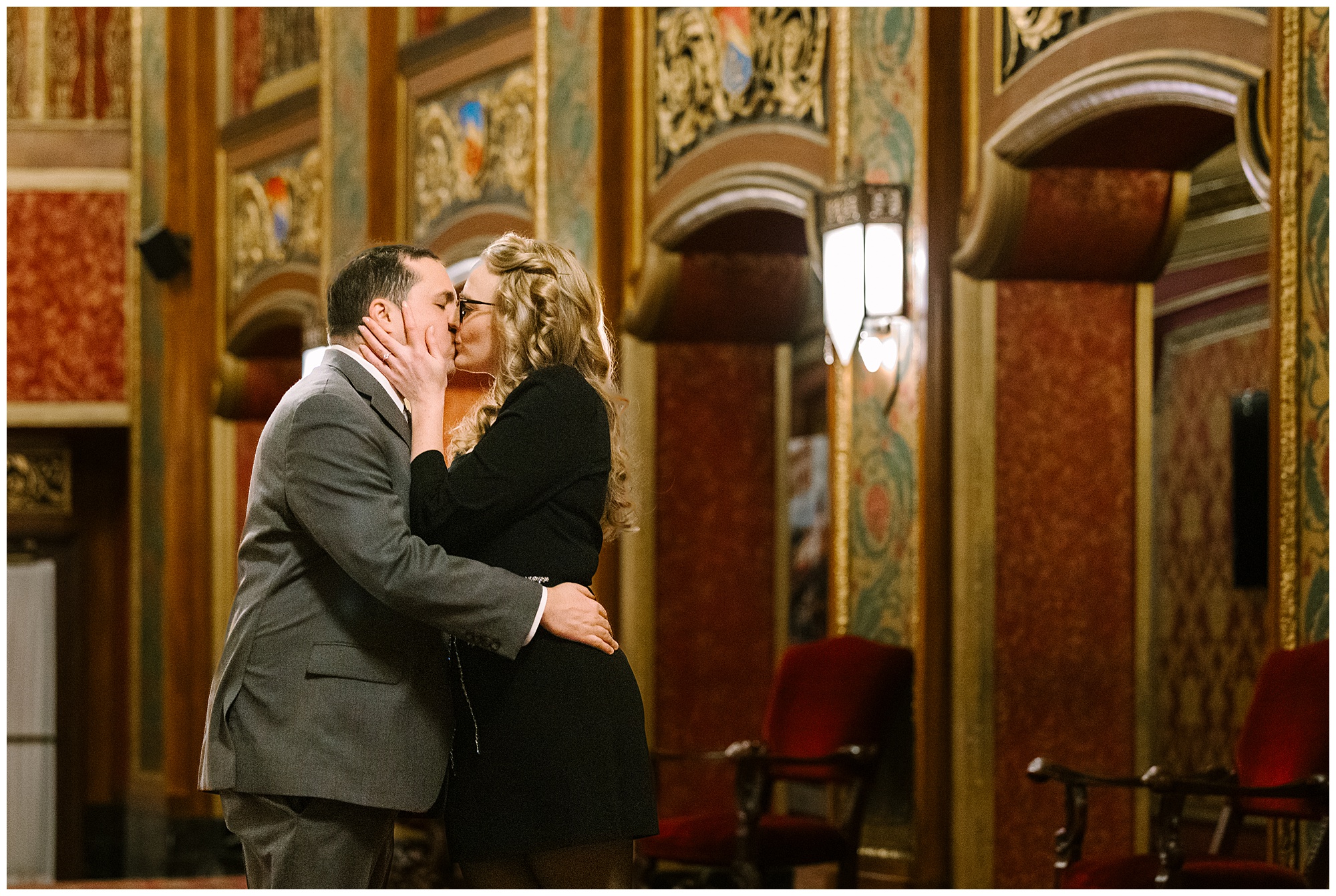 A couple dressed formally kisses in an ornate, vintage-style theater with red cushioned chairs and intricate gold and red wall designs in the background.