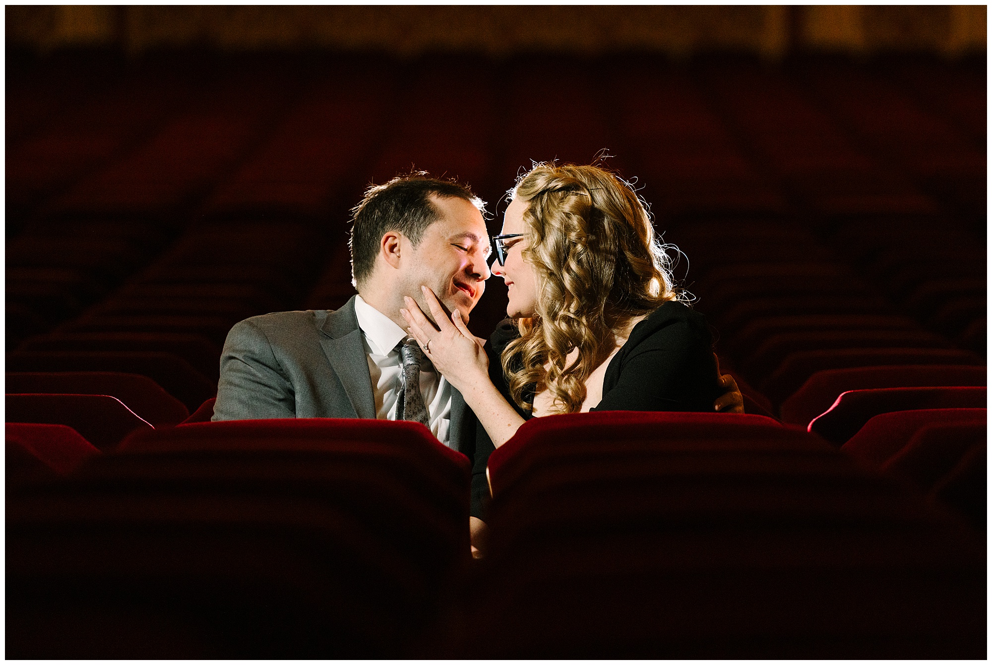A couple sits closely in a theater with red seats, gazing into each other’s eyes and smiling tenderly. The woman touches the man’s face gently, and a spotlight illuminates them in the otherwise dark auditorium.