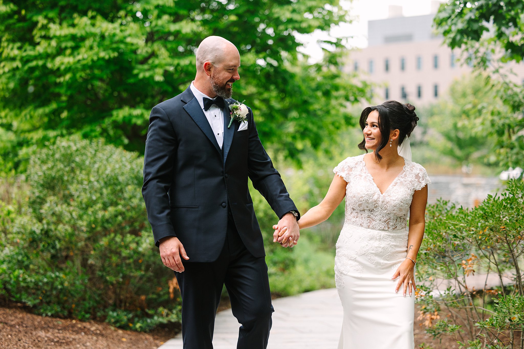 A couple dressed in wedding attire smiles at each other and holds hands while walking outdoors on a path surrounded by greenery, capturing the joy of their Williams Inn wedding.
