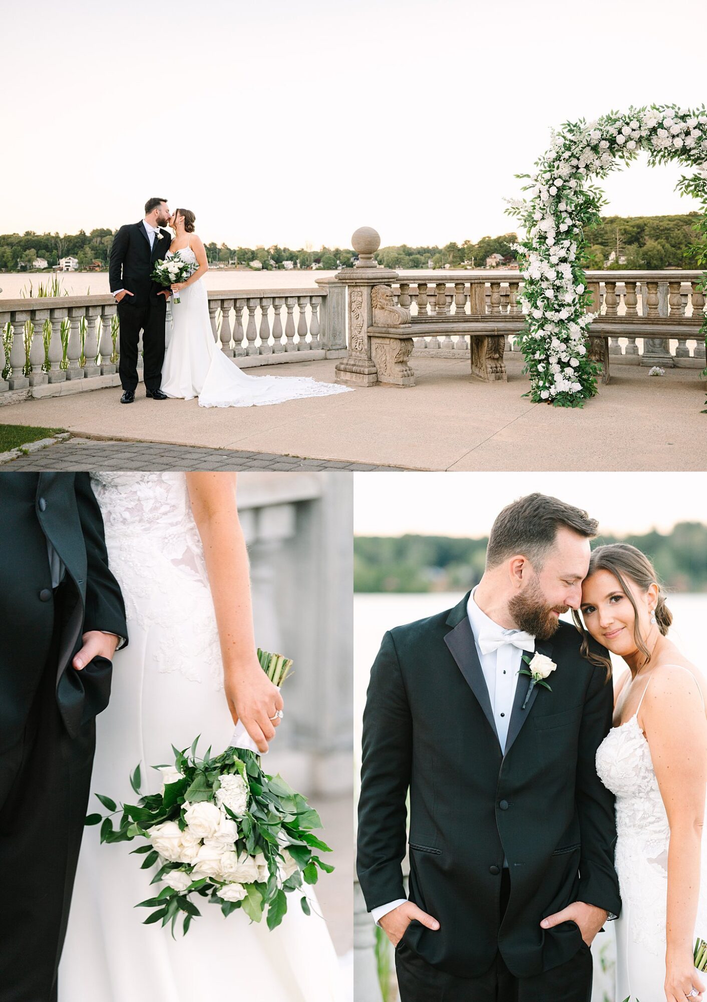 A bride and groom pose by a lakeside stone railing with a floral arch. The bride holds a bouquet of white flowers. They smile and lean affectionately, dressed in classic wedding attire.