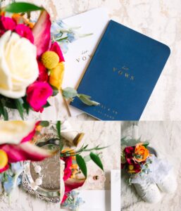 A flat lay of wedding vow books, a colorful bouquet, a silver tray with rings, a white rose boutonniere, and white shoes decorated with flowers on a marble surface.