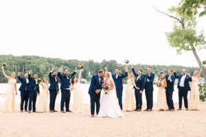 A bride and groom kiss on a sandy shore while their wedding party, dressed in navy suits and light dresses, cheer with raised bouquets near a lake and trees.