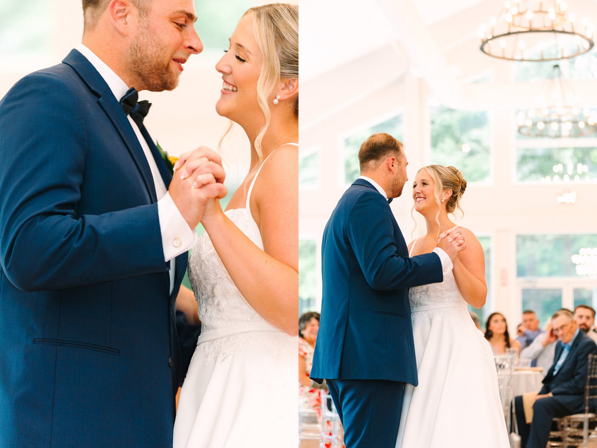A bride and groom smile and hold hands while sharing their first dance at a wedding reception, surrounded by seated guests in a bright, elegant venue.