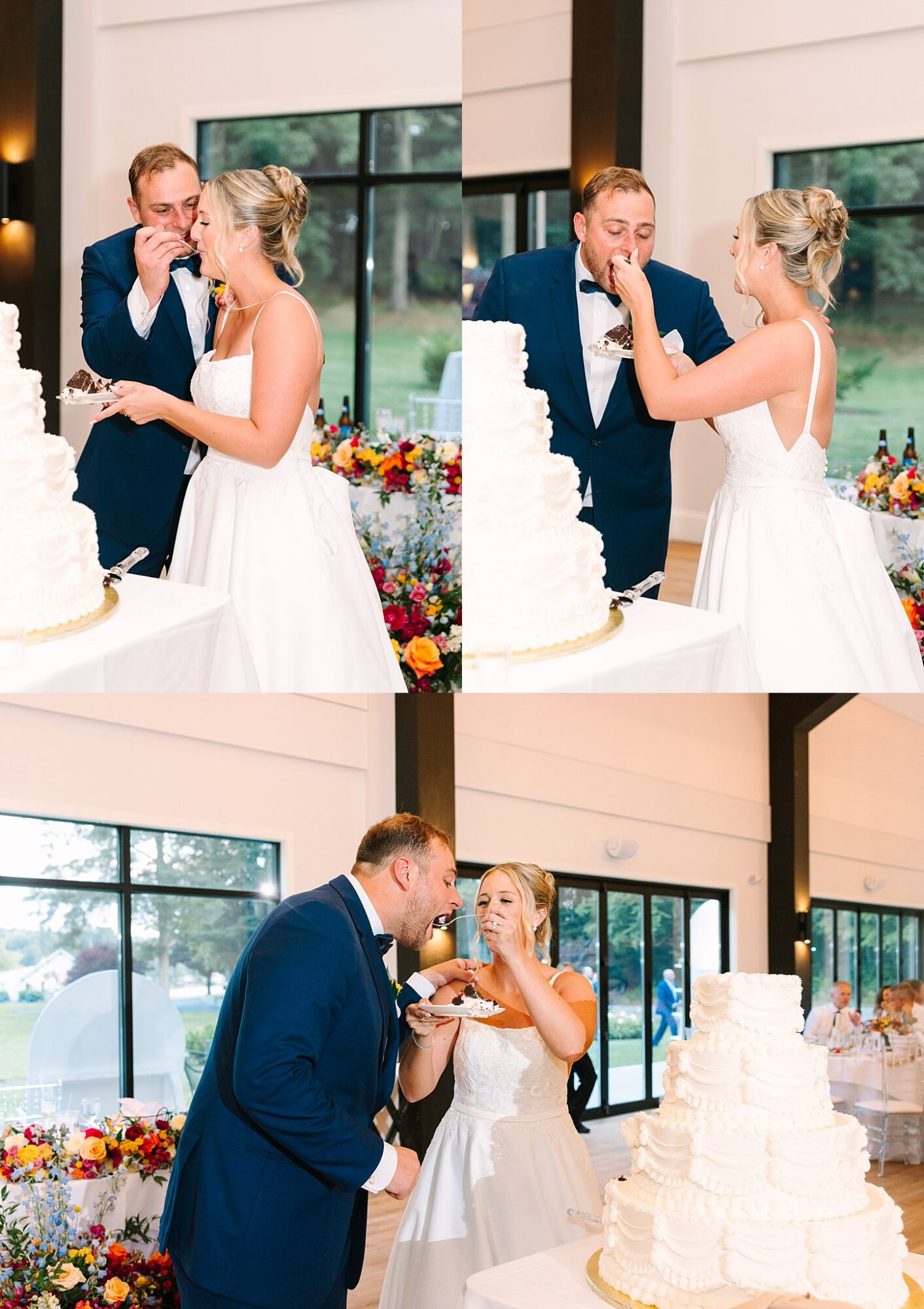A bride and groom feed each other wedding cake in a bright room with large windows and colorful flowers, both smiling and laughing during the celebration.