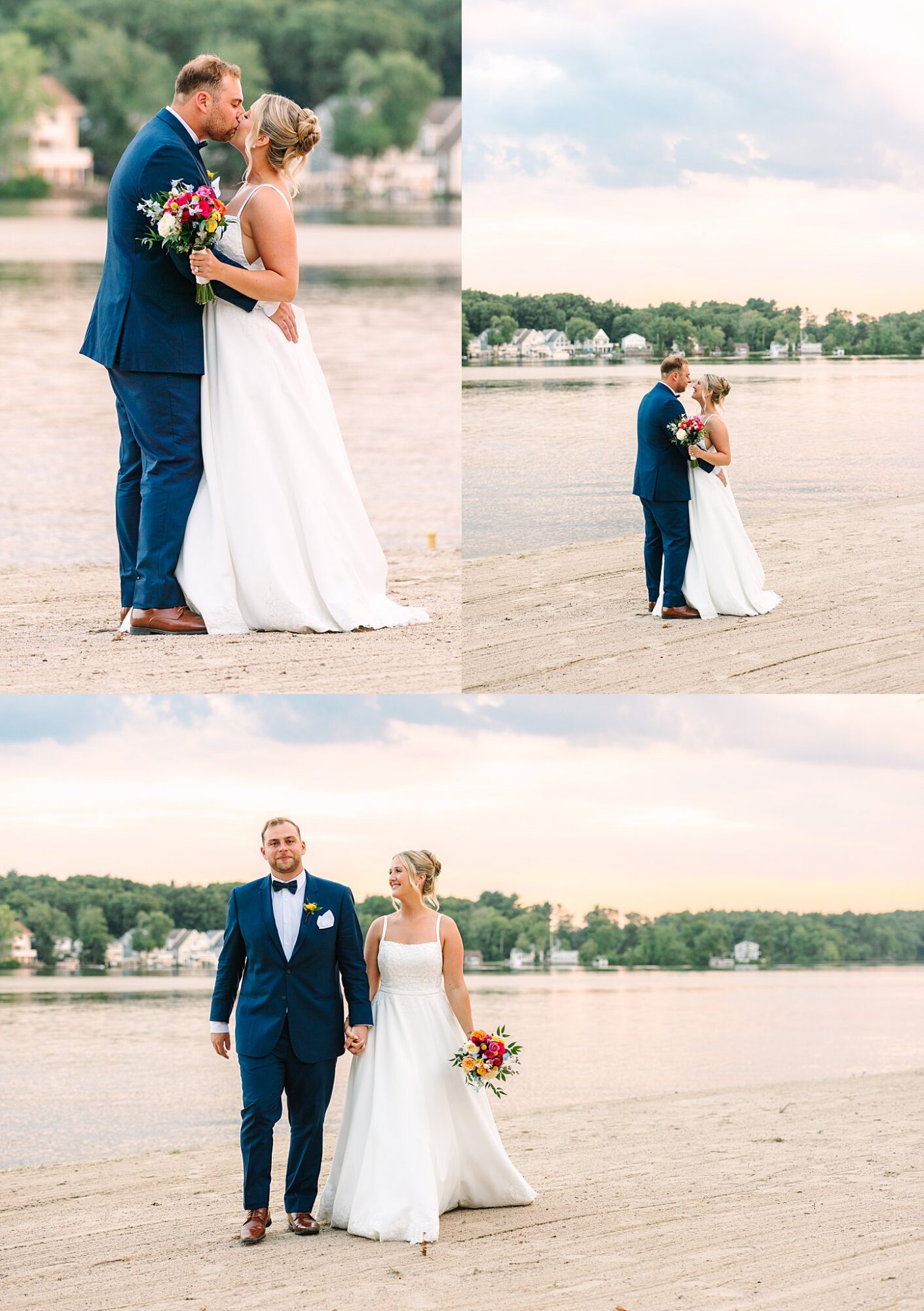 A bride and groom in wedding attire walk and embrace on a sandy beach by the water, holding a bouquet, with houses and trees in the background under a cloudy sky.