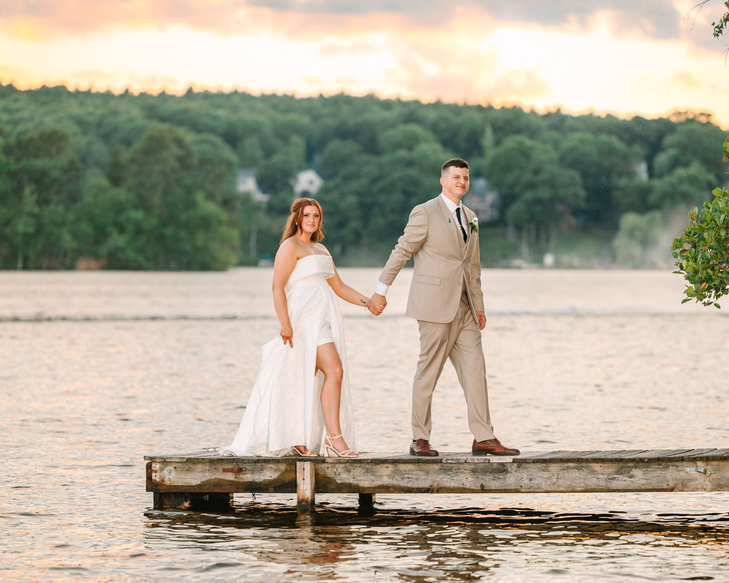 A bride and groom hold hands, standing on a wooden dock by a lake at sunset, surrounded by trees and calm water—capturing the romance of a Lake Pearl Wrentham Wedding.