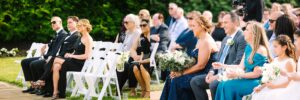 Wedding guests sit outdoors on white chairs, dressed formally. Some hold bouquets, and everyone faces forward, watching the ceremony attentively on a sunny day. Greenery is visible in the background.