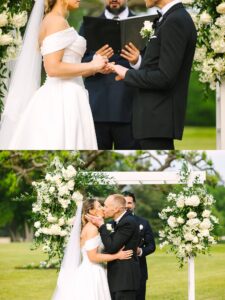 A bride and groom exchange rings, then share a kiss at their outdoor wedding ceremony, standing in front of a floral arch with a person officiating.