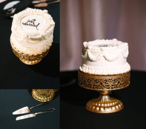 A small white wedding cake with just married written on top sits on an ornate gold stand. Nearby are a cake knife and server placed on a dark tablecloth.