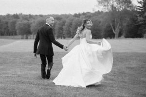 A bride and groom walk hand in hand across a grassy field, smiling back at the camera. The brides long dress flows behind her, and trees are visible in the background. The photo is in black and white.