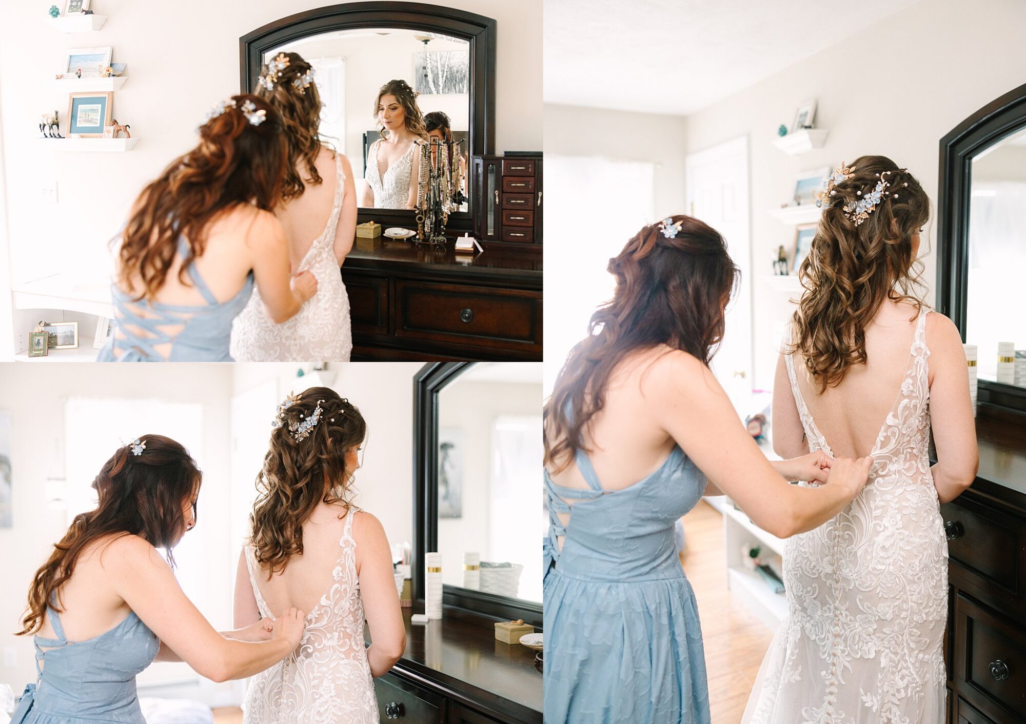 A woman in a blue dress helps another woman in a white lace wedding gown zip up her dress in a bright room with a dark wooden dresser and mirror.