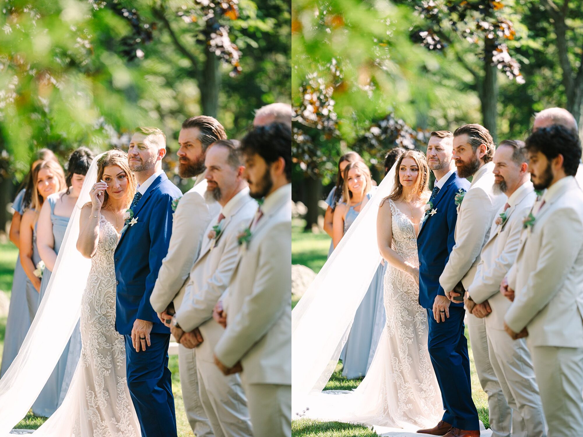 A bride and groom stand outdoors with their wedding party during a ceremony. The groom wears a blue suit, and the bride in a white dress wipes her eye. Bridesmaids and groomsmen stand nearby in light-colored outfits.