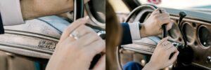 Two peoples hands, both wearing rings, grip the gearshift inside an old car. The dashboard and part of the steering wheel are visible, with a Chevelle by Chevrolet emblem seen on the left.