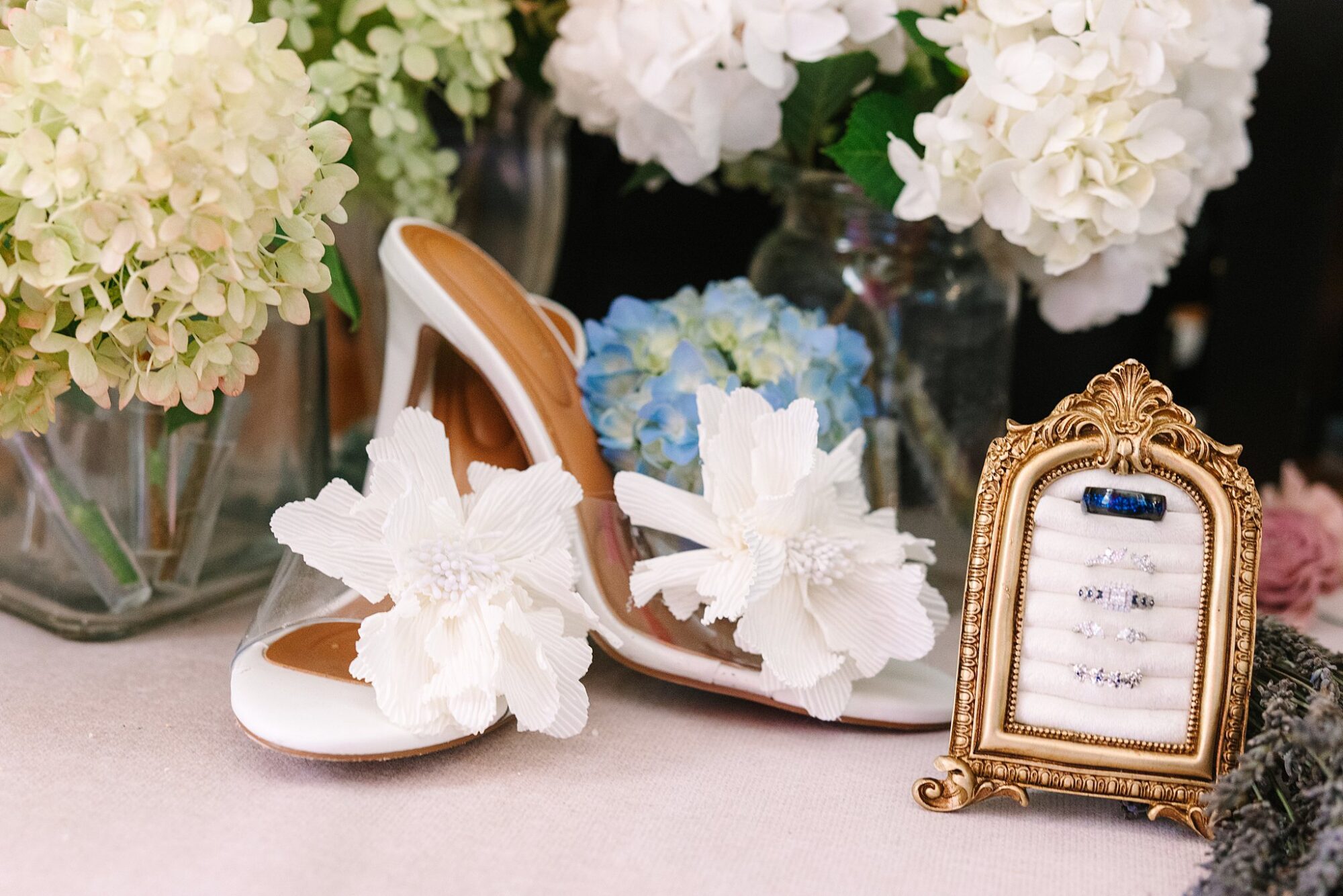 White heels with large floral decorations are displayed among vases of white and green hydrangeas. A gold ornate frame holds a set of rings on display beside the shoes. The setting appears elegant and romantic.