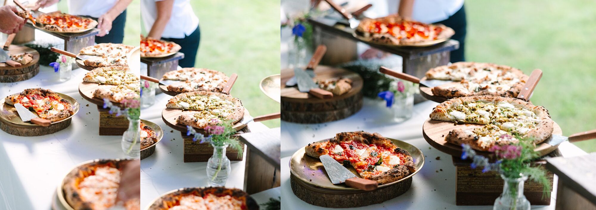 A variety of fresh pizzas on wooden stands are displayed on a white table outdoors, with people serving slices and small vases of flowers as decoration.