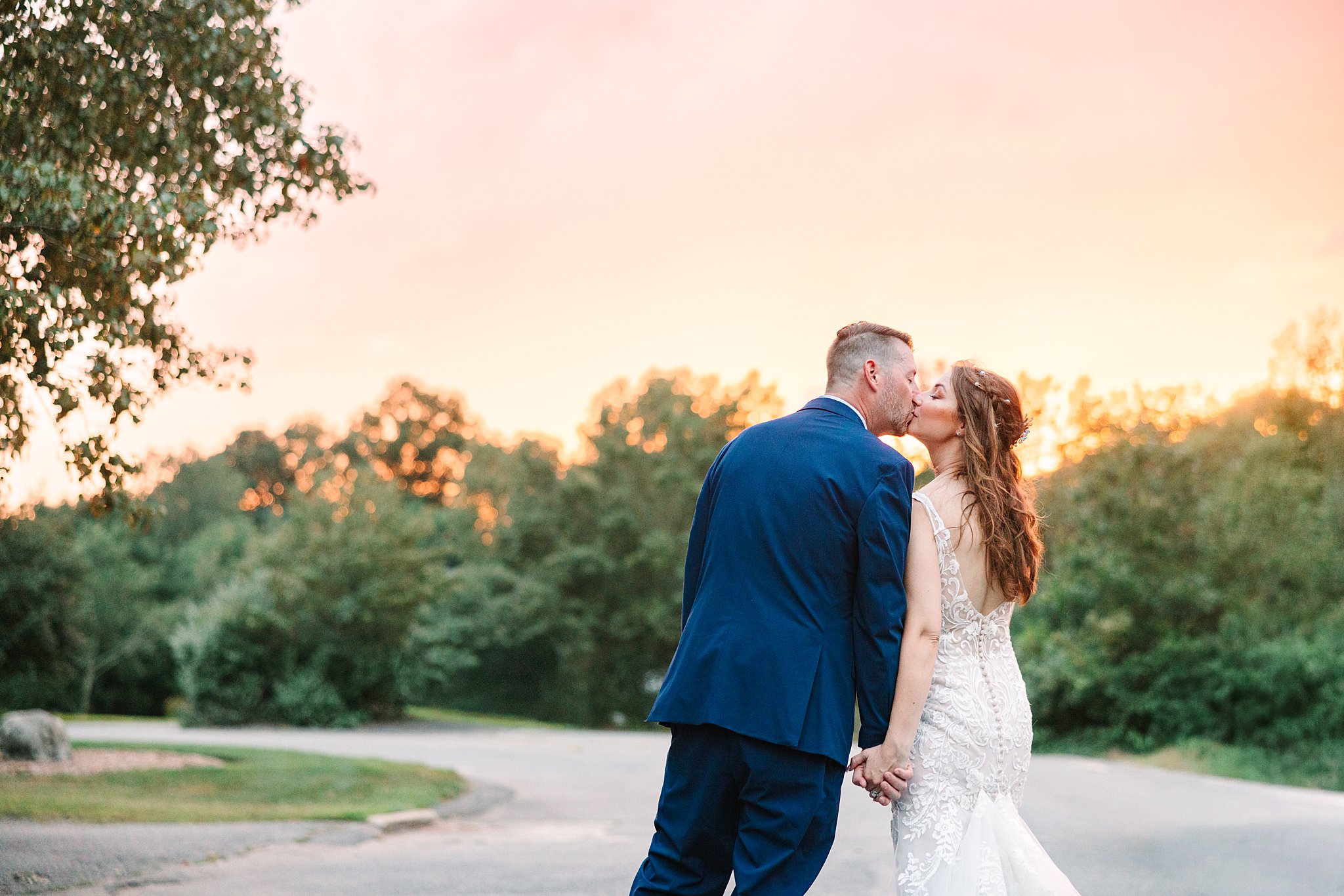 A bride and groom hold hands and kiss on a road at sunset, with trees and a colorful sky in the background.