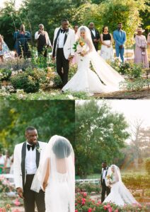 A bride in a white dress and a groom in a white jacket walk together outdoors through a flower garden, surrounded by smiling guests and lush greenery.