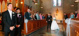 A bride in a white dress walks down the church aisle with an older man as guests watch. The groom stands at the altar, smiling, with groomsmen beside him. Stained glass windows and pews decorate the scene.