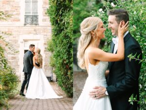 A bride in a white dress and a groom in a black suit smile and embrace outdoors near a stone building and greenery on their wedding day.