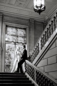 A bride and groom stand on a grand staircase, gazing at each other. Sunlight filters through a large window behind them. The photo is in black and white, with ornate architecture and a hanging light fixture above.