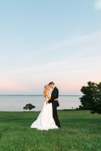 A bride and groom stand embracing on a grassy hill by the water at sunset, with a light pink and blue sky above them and trees in the background.