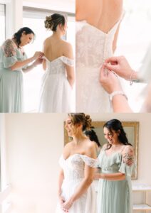 A bridesmaid in a sage green dress helps a bride in an off-shoulder white gown button up her dress. Both women are smiling in a bright, softly lit room.