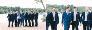 Groomsmen in navy suits and a young boy in suspenders stand on a sandy beach by a lake; in the second photo, four men in suits walk together, smiling. Trees and houses are visible in the background.