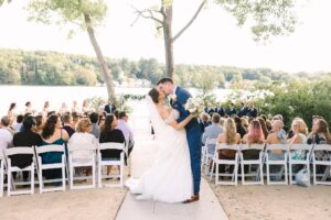 A bride and groom share a kiss at an outdoor wedding ceremony by a lake, surrounded by guests seated on white chairs, with trees and water in the background.
