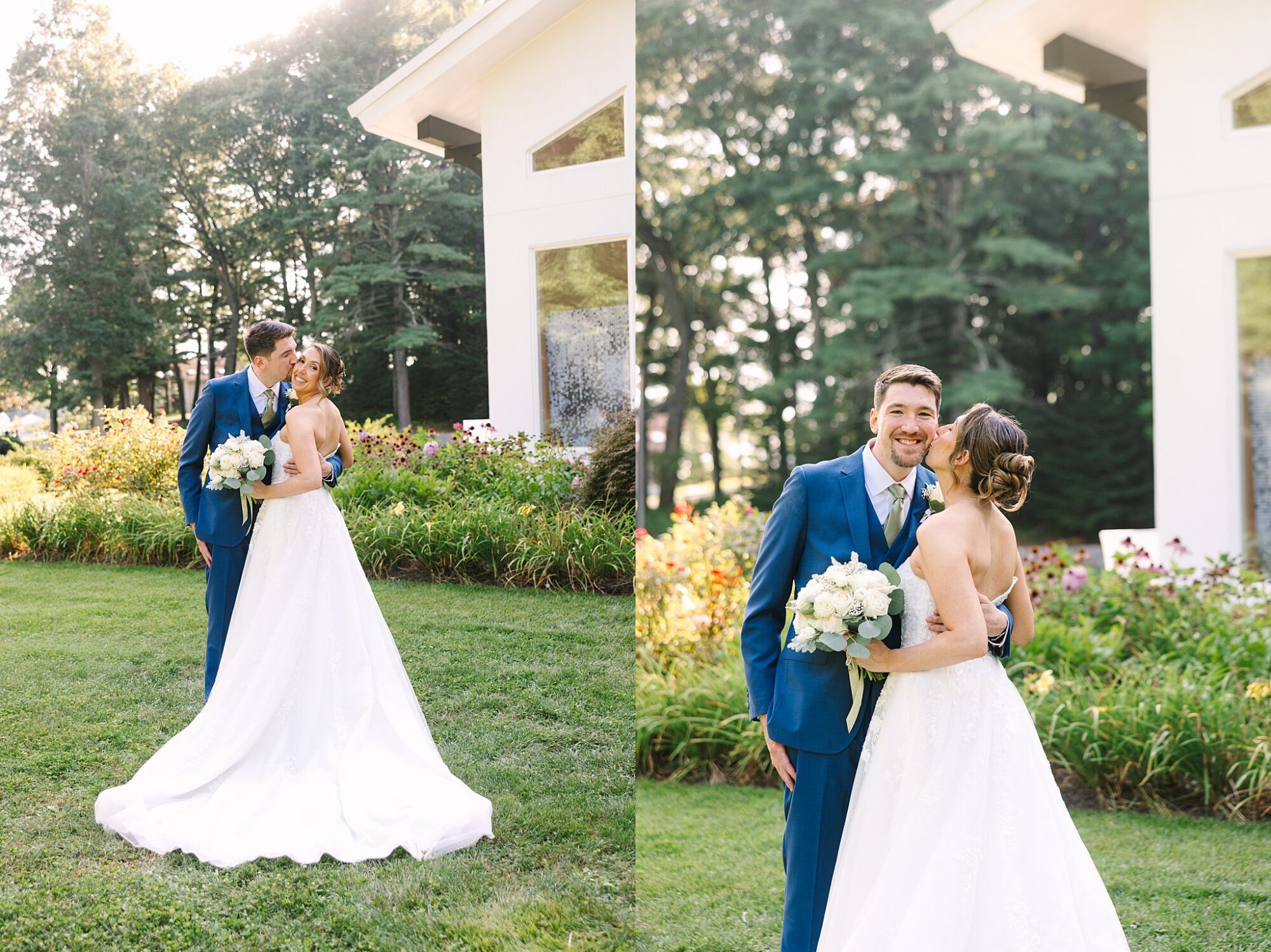 A bride in a white gown and a groom in a blue suit smile and embrace outdoors near a white building surrounded by greenery and flowers.