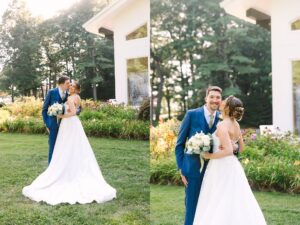A bride in a white gown and a groom in a blue suit smile and embrace outdoors near a white building surrounded by greenery and flowers.