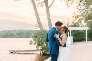 A bride and groom kiss near a lake at sunset, with trees, a dock, and a white pergola in the background. The bride wears a white dress; the groom wears a blue suit.