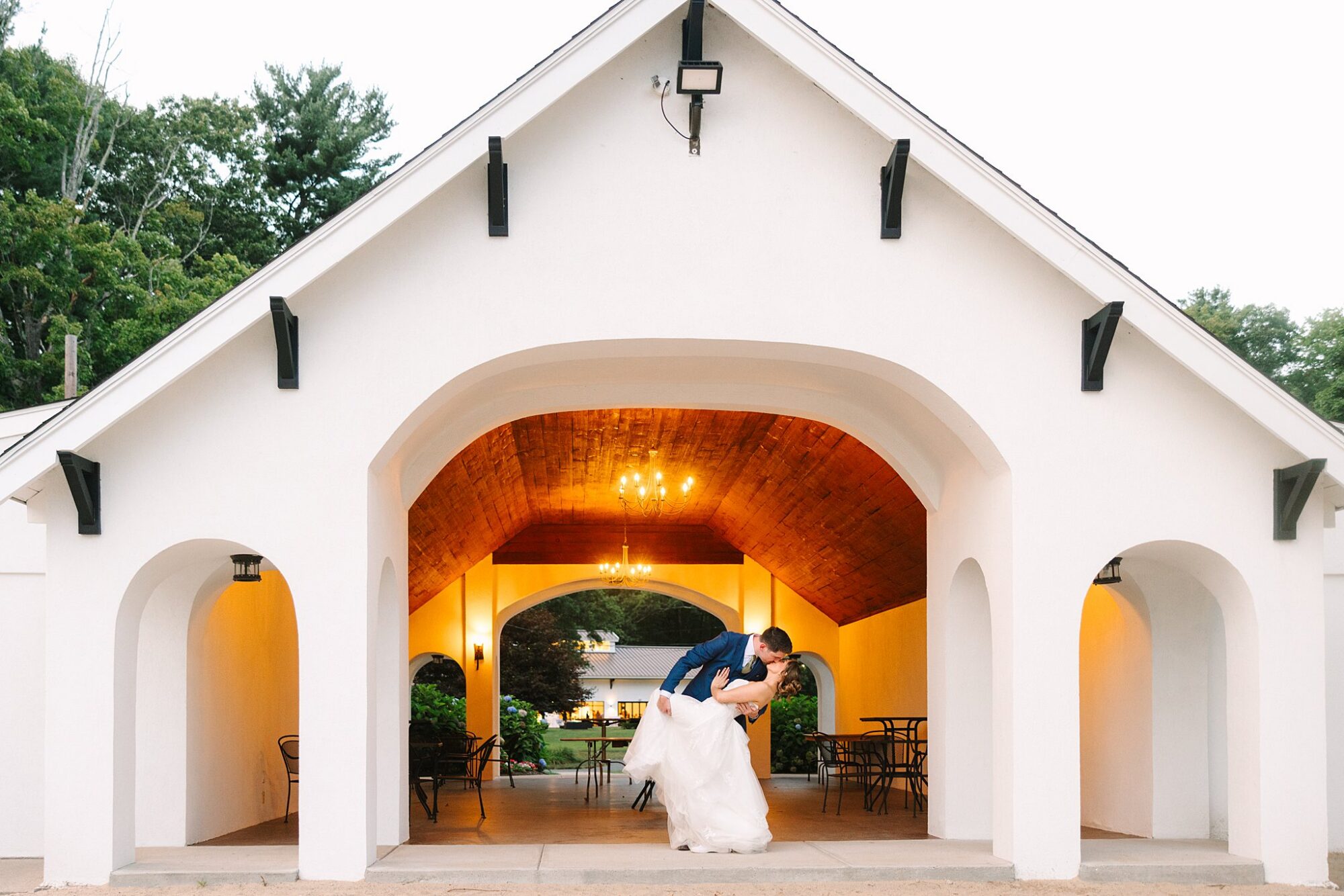 A bride and groom share a kiss as the groom dips the bride under a white pavilion with arches and warm lights, surrounded by trees.